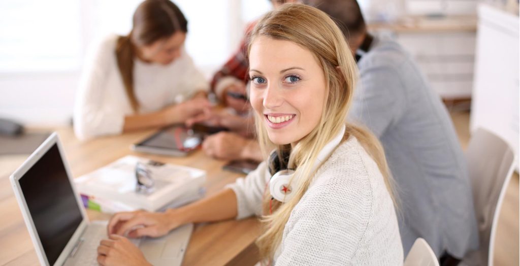 A blonde woman sits at a desk with a laptop in Braunschweig. In the background, 3 other people sit with mobile phones.